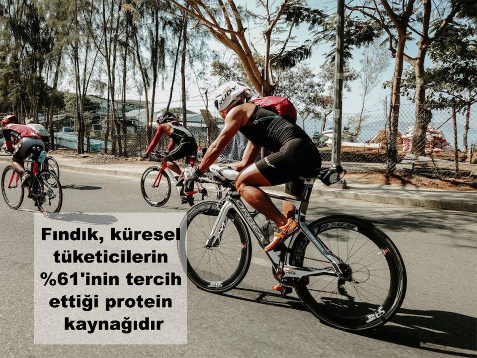 Ağaçlarla çevrili bir caddede yarışan bisikletçiler. Metin: Kuruyemişler küresel tüketicilerin %61'i için tercih edilen protein kaynağı